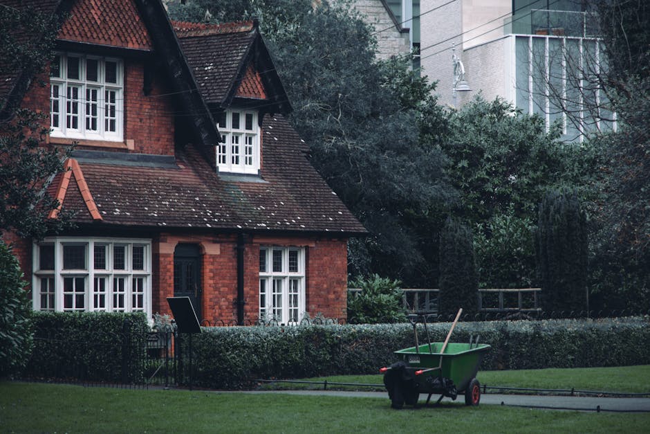 A traditional red-brick house with a steep, tiled roof and white-framed windows situated in a well-maintained garden. The garden features a neatly trimmed hedge bordering a green lawn, with a pathway 