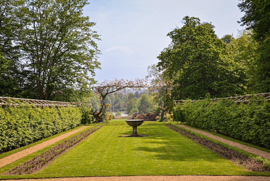 A peaceful park scene featuring a wide, straight gravel pathway lined with small, evenly spaced trees that have lush green leaves. On either side of the path, there are garden beds filled with vibrant